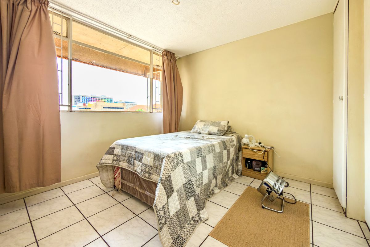 A bedroom featuring tiled flooring, a built-in cupboard, and a large window with security bars, offering natural light and city views.