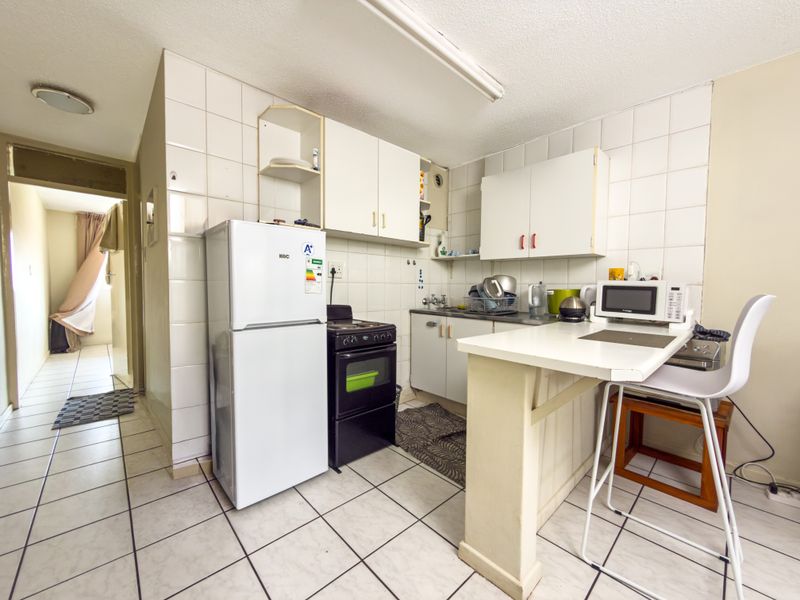 A compact kitchen featuring tiled flooring, white cabinetry, a breakfast bar, and a tiled splashback, offering a functional layout in need of cosmetic updates.