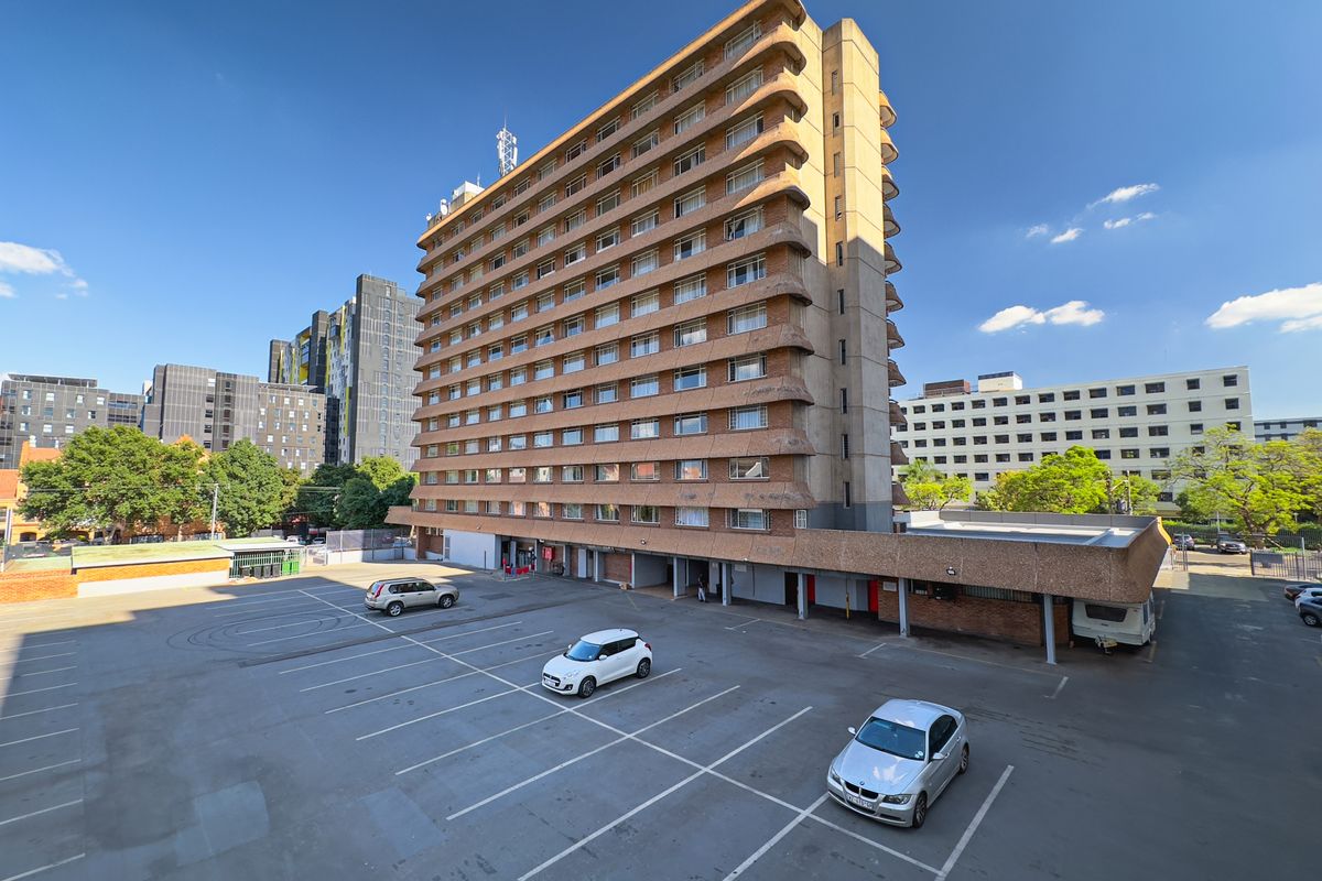 Exterior view of a multi-story apartment building featuring a large asphalt parking area and concrete facade with balconies.