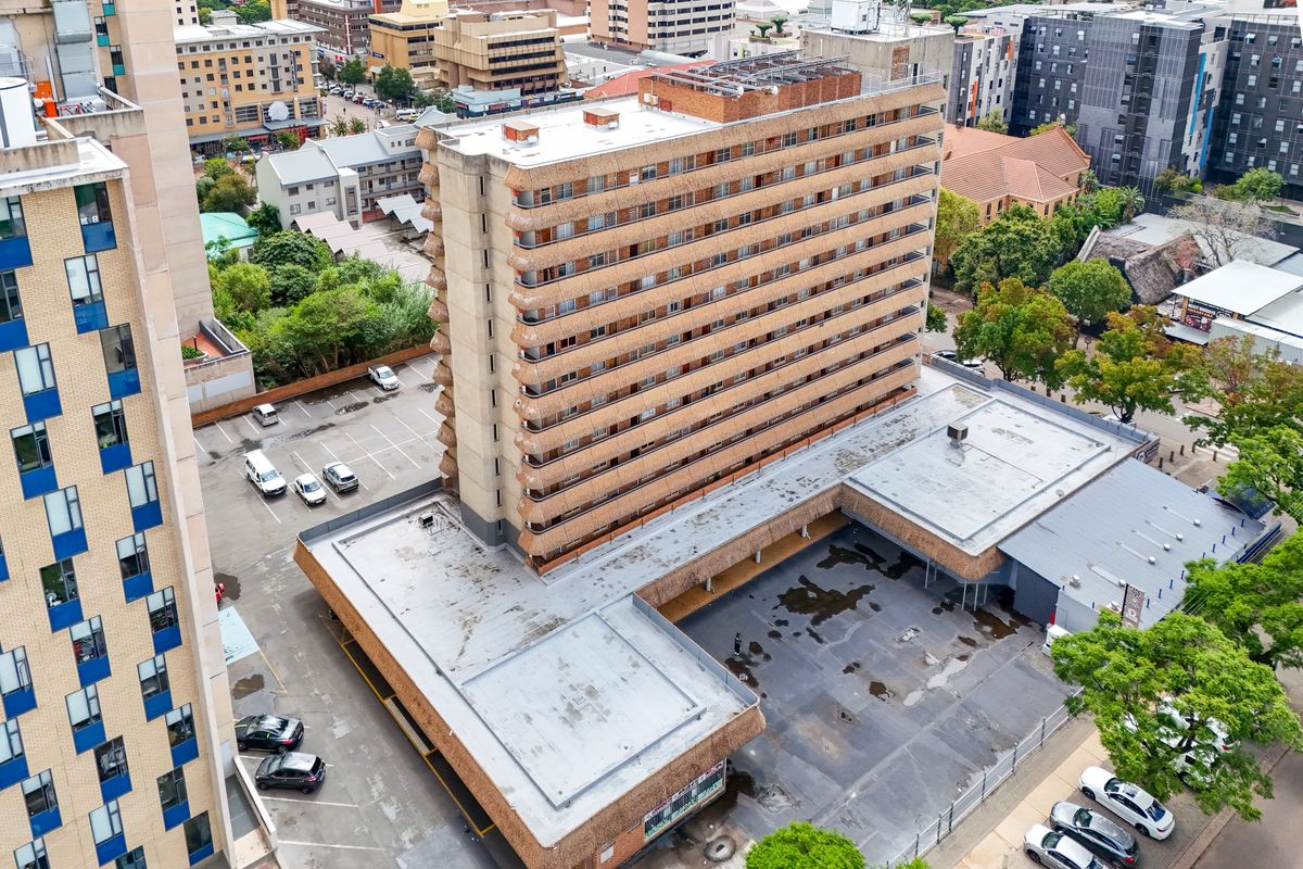 Aerial view of a multi-story apartment building featuring a flat roof, private balconies, and an adjacent paved parking lot in an urban setting.