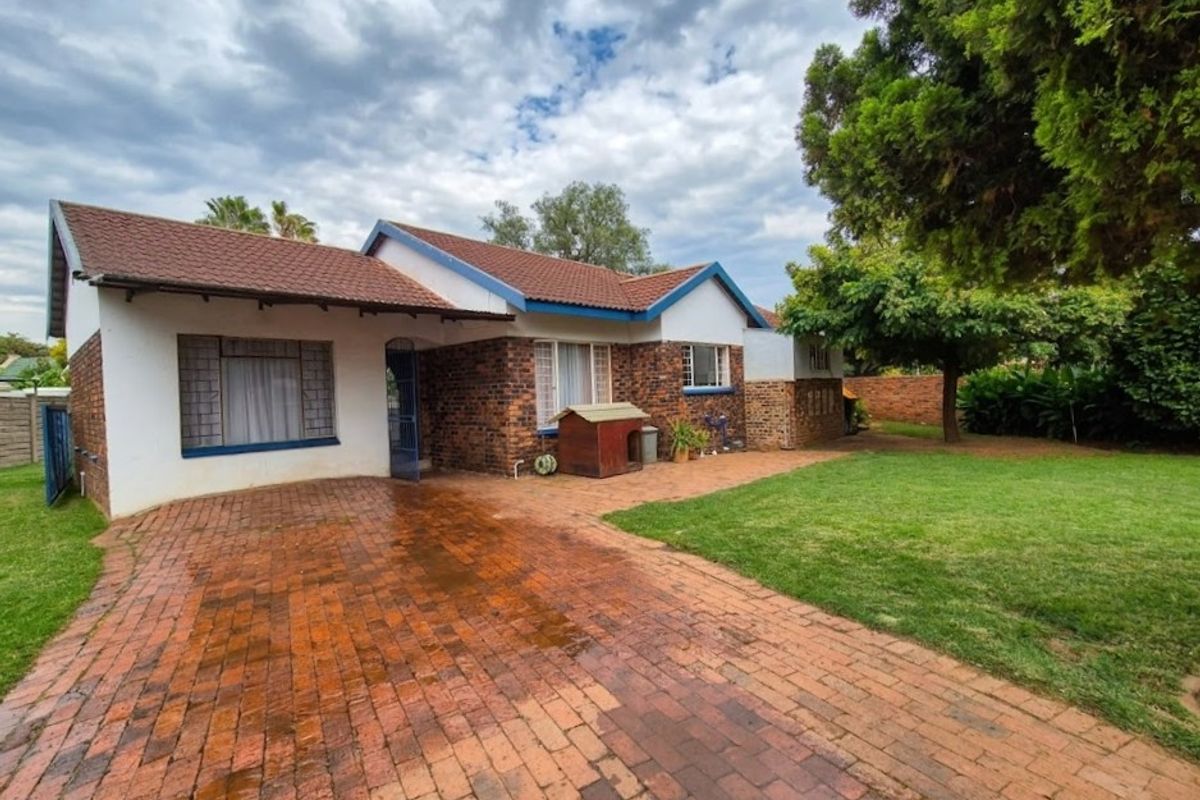 Rear view of a single-story brick house featuring a paved driveway, tiled roof, and a spacious garden area.