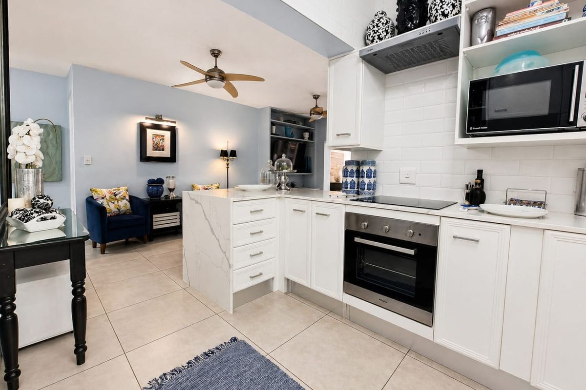 An open-plan kitchen and living area featuring modern white cabinetry, stone countertops, integrated appliances, and neutral tiled flooring.