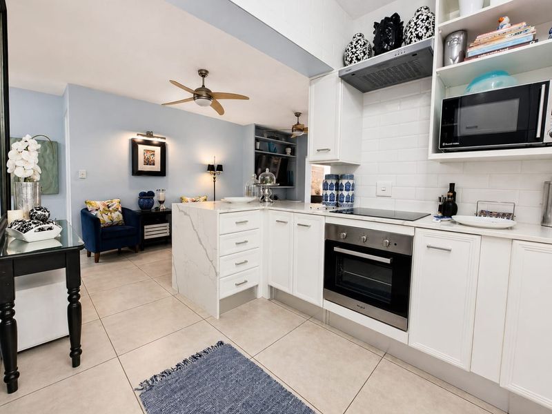 An open-plan kitchen and living area featuring modern white cabinetry, stone countertops, integrated appliances, and neutral tiled flooring.