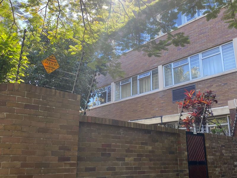 Front exterior view of a brick apartment building featuring security electric fencing and established boundary walling.