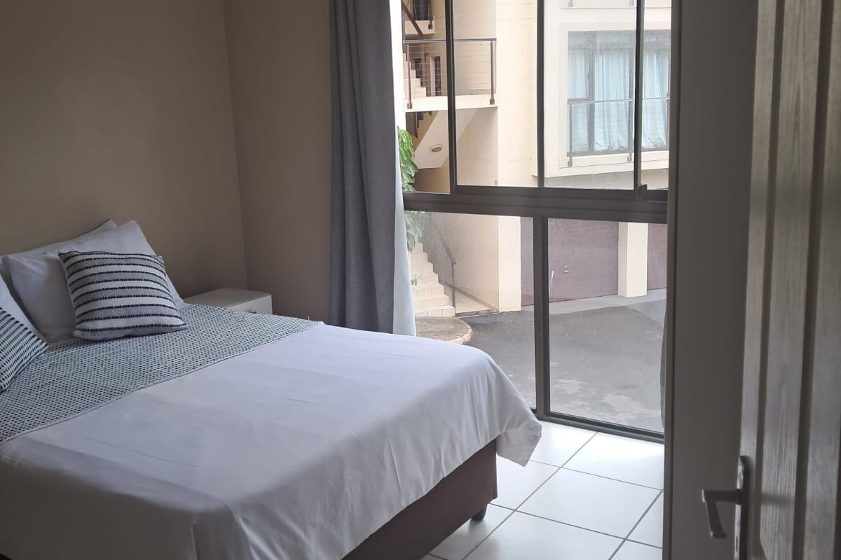 A bedroom featuring tiled flooring, a ceiling fan, and large sliding glass windows providing natural light and views of the complex exterior.