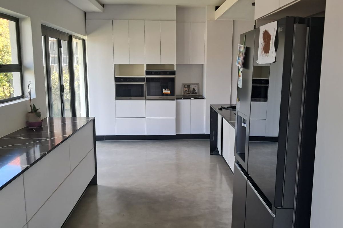 Modern kitchen featuring sleek white handleless cabinetry, dark stone countertops, integrated appliances, and polished concrete flooring with ample natural light.