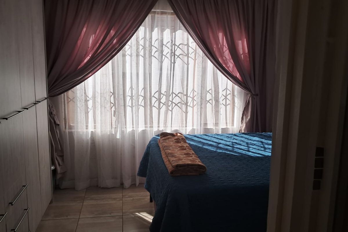 A bedroom featuring built-in cupboards and tiled flooring, with natural light filtering through a window protected by security bars.