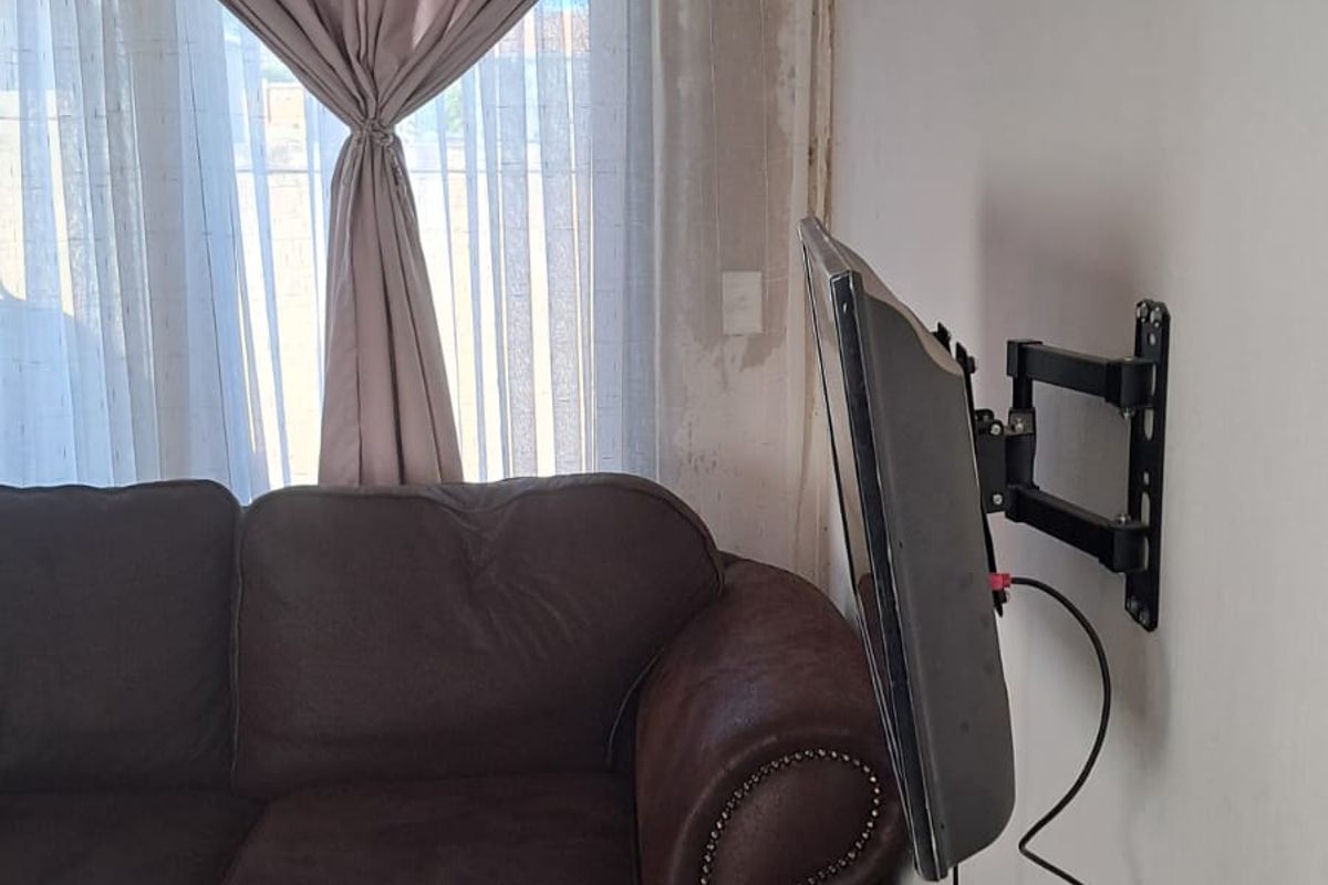 A living room area featuring a wall-mounted television bracket and tiled flooring, showing signs of wall surface deterioration near the window.