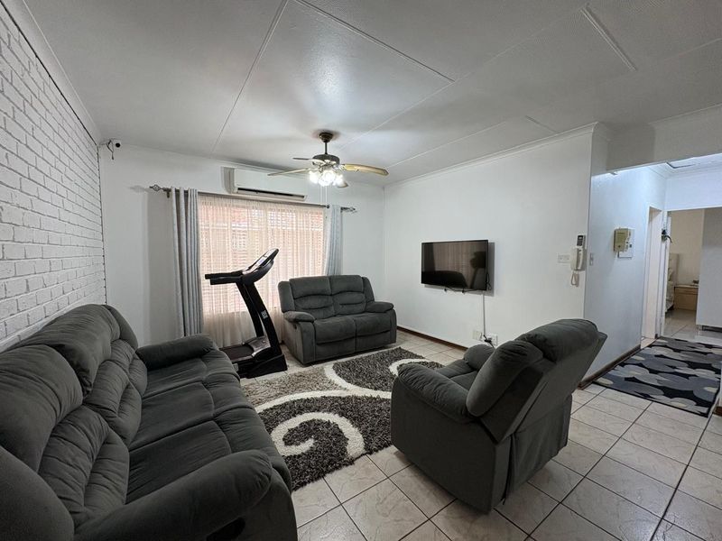 A comfortable living room featuring tiled flooring, an exposed brick feature wall, a ceiling fan, and a wall-mounted split-system air conditioner.