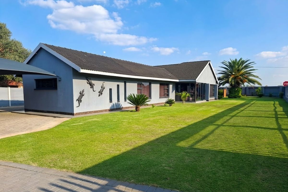 A well-maintained single-story house featuring a tiled roof, paved driveway with carport, and a spacious, manicured lawn with perimeter fencing.