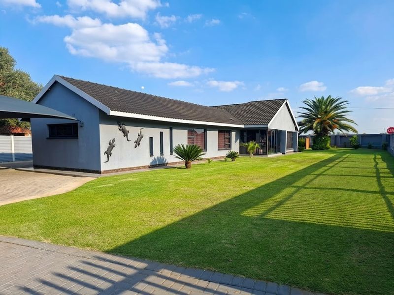 A well-maintained single-story house featuring a tiled roof, paved driveway with carport, and a spacious, manicured lawn with perimeter fencing.