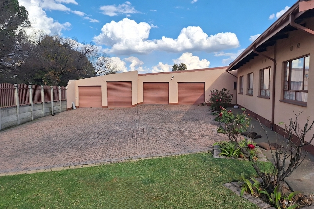 Spacious rear exterior area featuring a large paved driveway and a four-car garage with metal roller doors, set against a well-maintained garden.