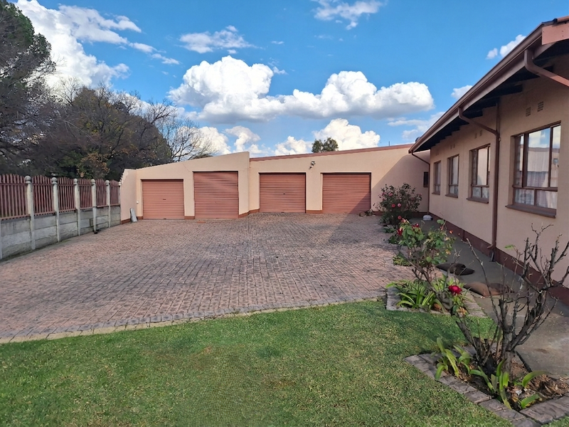 Spacious rear exterior area featuring a large paved driveway and a four-car garage with metal roller doors, set against a well-maintained garden.