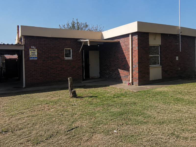 A single-story brick house featuring a side carport, roller garage door, and security bars on windows, set on a large grassed plot.