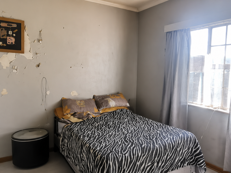 A bedroom featuring a window with curtain rails, currently showing significant wall surface damage and peeling paint requiring repair and redecoration.
