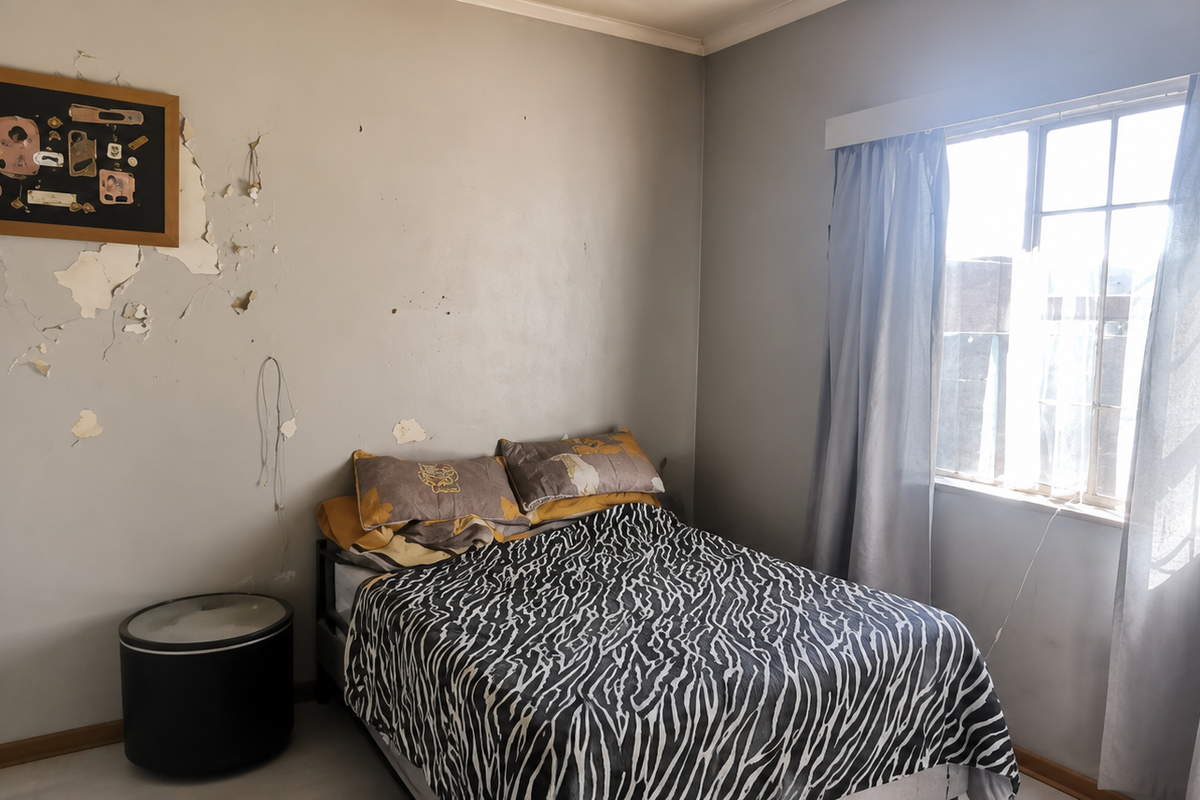A bedroom featuring a window with curtain rails, currently showing significant wall surface damage and peeling paint requiring repair and redecoration.