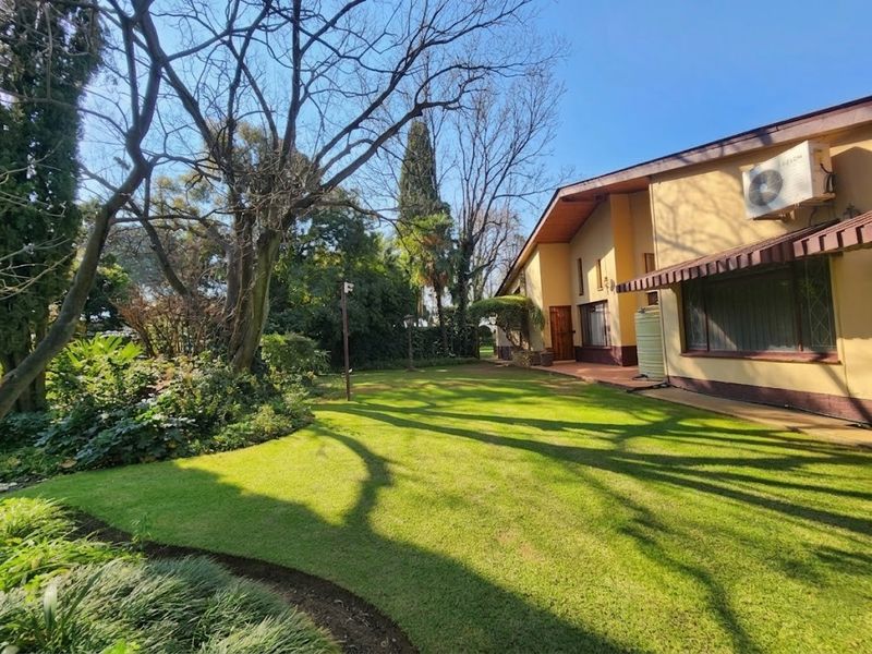 A spacious rear garden featuring a well-maintained lawn, mature trees, and a covered patio area attached to the main house structure.