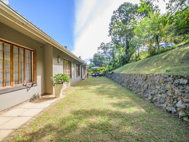 A well-maintained rear garden featuring a stone retaining wall, a paved walkway along the house, and established trees on a sloped embankment.