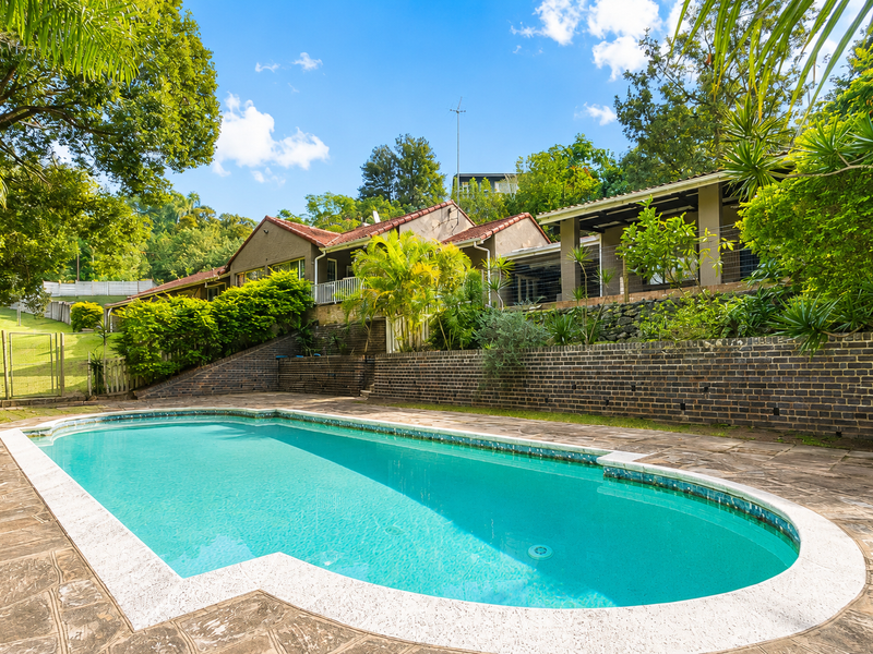 A well-maintained swimming pool area featuring stone paving, a brick retaining wall, and a covered patio overlooking the garden.