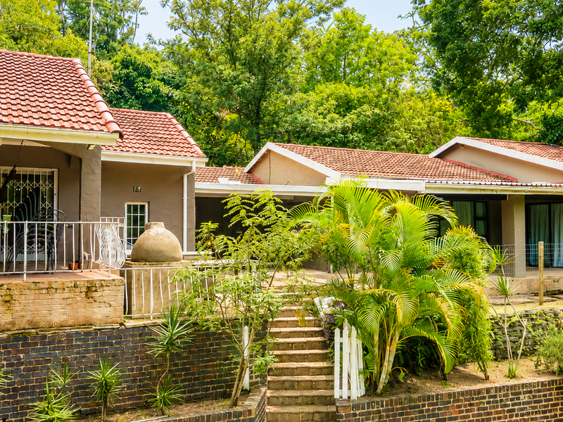 Rear view of the property featuring a covered patio, stone staircase, built-in pizza oven, and tiered brick retaining walls set against a lush garden.
