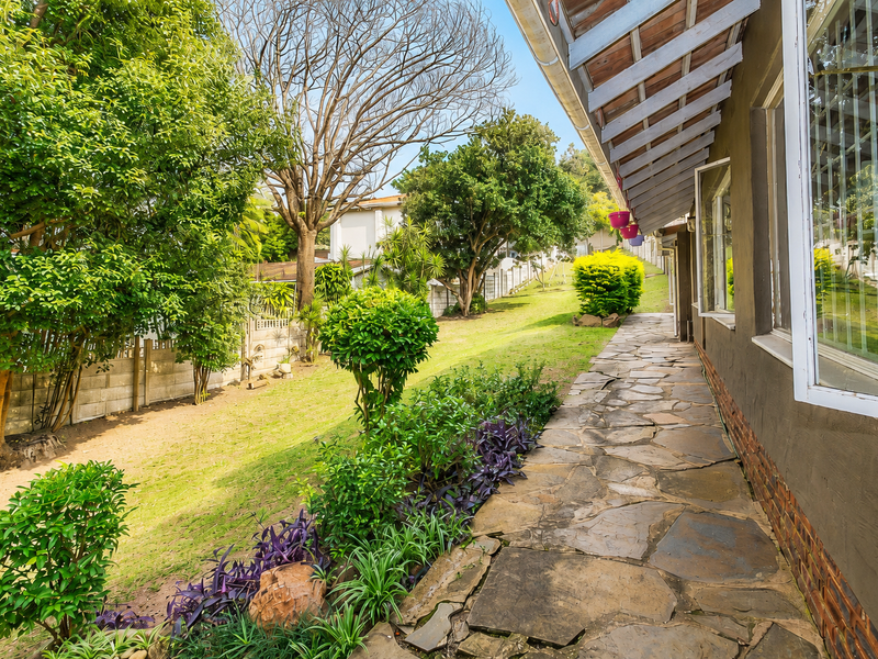 A rear exterior view featuring a stone paved walkway along the house, mature garden landscaping, and a precast concrete boundary wall.