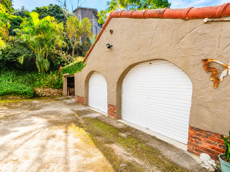 A double garage with white roller doors, textured stucco finish, and terracotta roof tiles, situated on a concrete driveway with surrounding garden greenery.