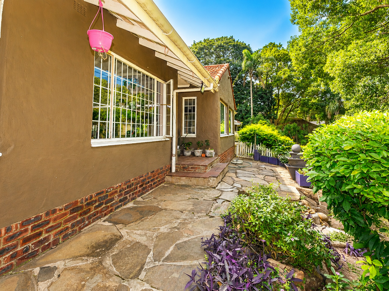 Rear exterior view of the house featuring a stone paved patio, brick base detailing, and established garden surroundings.