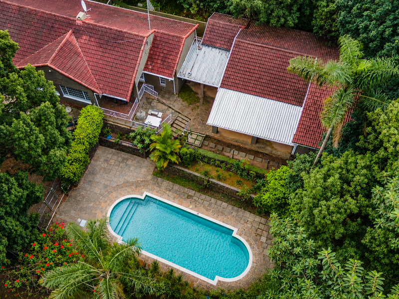 Aerial view of a residential property featuring a swimming pool, paved patio area, and mature garden surroundings with tiled and metal roofing.