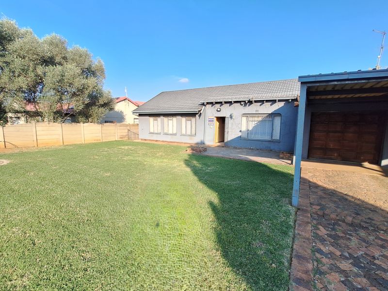 Rear view of the property featuring a spacious lawn, paved driveway, carport, and a tiled roof structure with security bars on windows.
