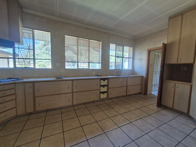 A spacious, empty kitchen featuring extensive built-in cabinetry, tiled flooring, a stainless steel sink, and large windows with security bars.