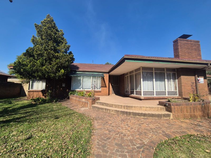 Front exterior view of a single-story brick house featuring a paved patio area, brick chimney, and established garden surroundings.