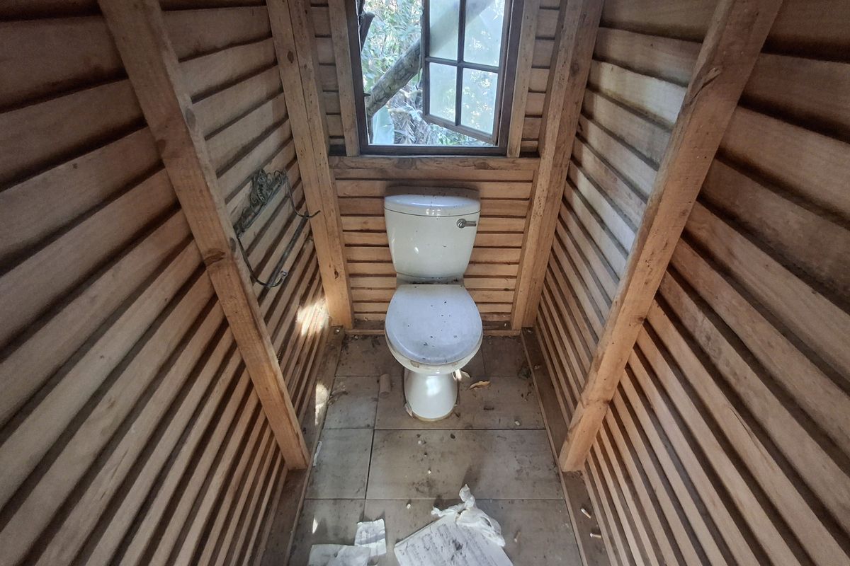 A rustic, timber-clad outdoor toilet facility featuring a basic porcelain toilet and a small window, requiring maintenance and refurbishment.