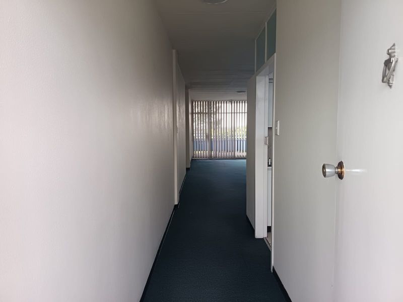 A long, empty hallway featuring dark carpet flooring and vertical blinds leading towards a bright, open living area.