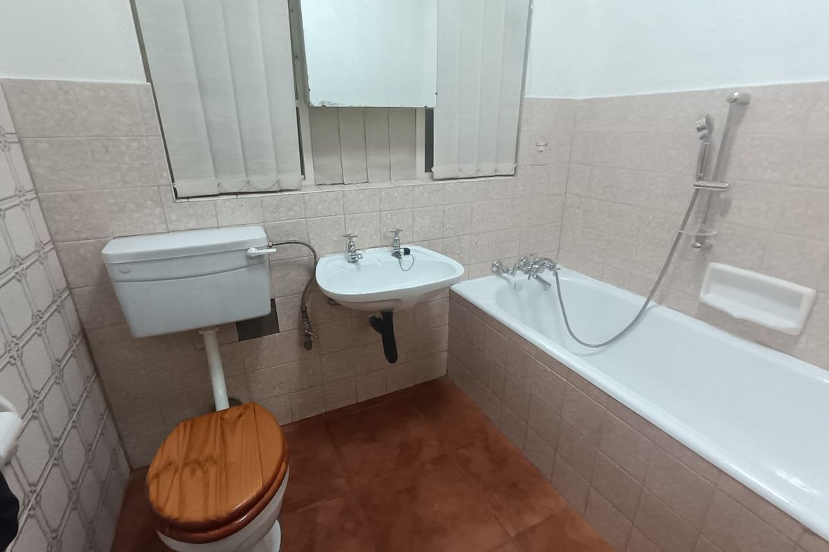 A functional bathroom featuring a bathtub with handheld shower, pedestal washbasin, and toilet, finished with neutral wall tiles and terracotta-style floor tiles.