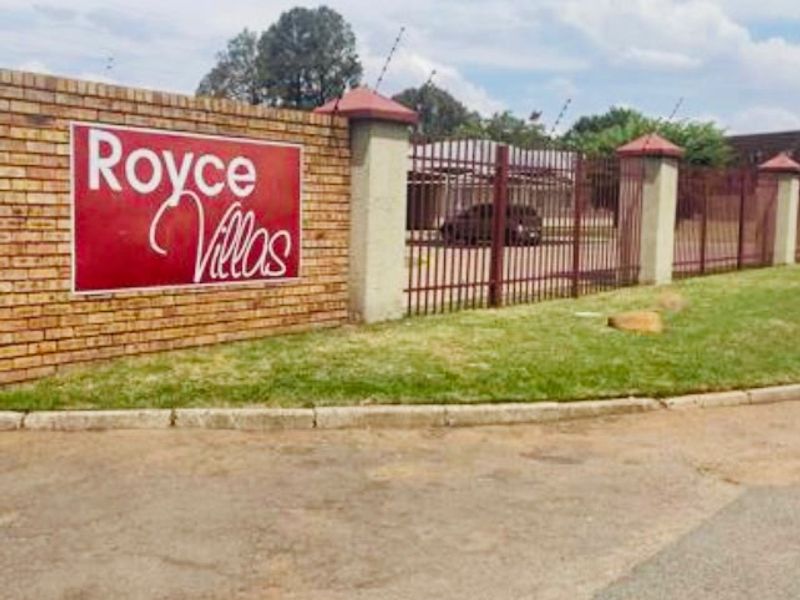 Street view of the Royce Villas complex entrance, featuring a brick boundary wall, security fencing, and gated access.