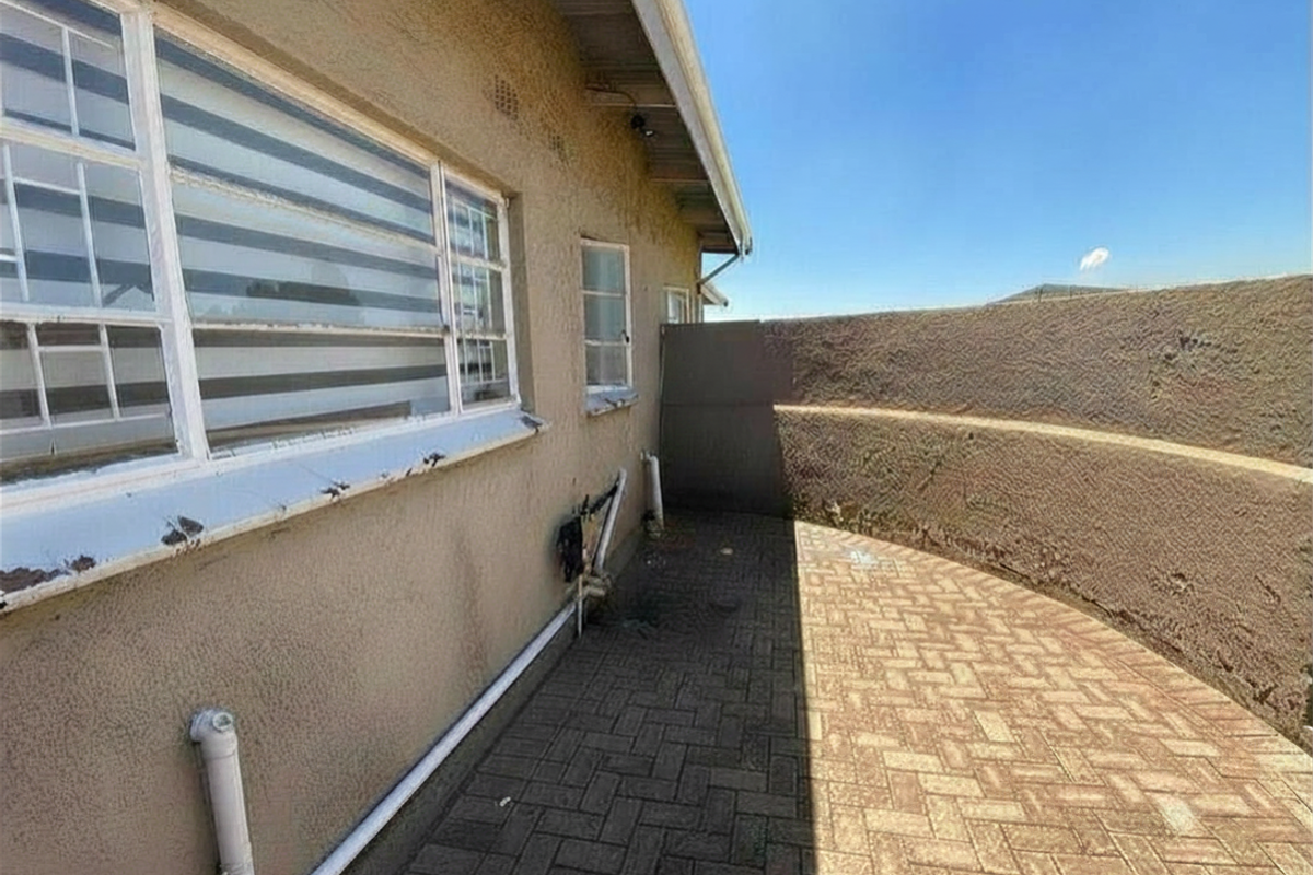 A narrow rear exterior walkway featuring paved flooring, a curved retaining wall, and external plumbing fixtures along the building facade.