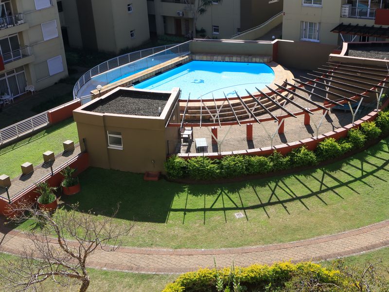 Aerial view of a communal apartment complex pool area featuring a swimming pool, pergola structure, and landscaped garden surrounds.