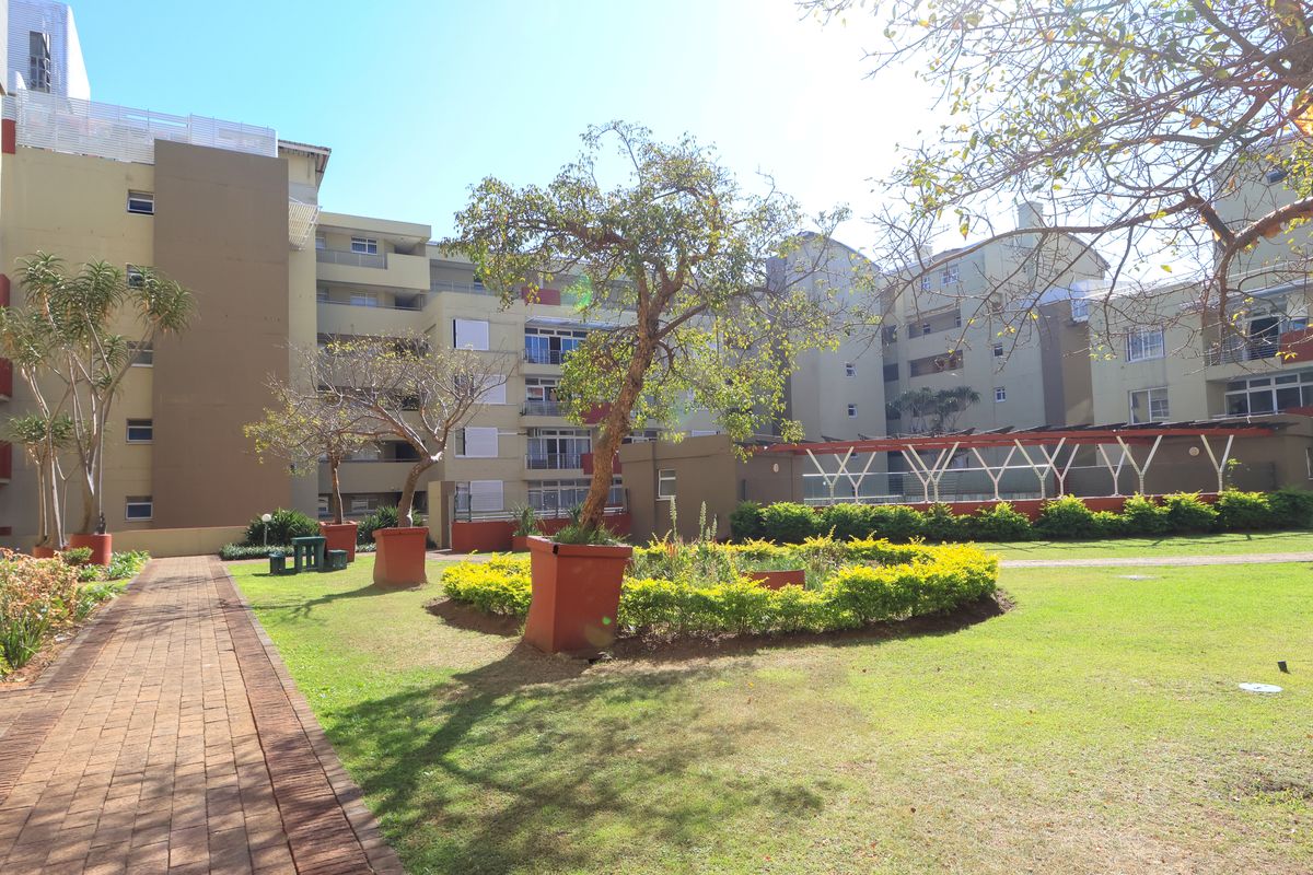 A well-maintained communal garden area featuring a paved walkway, manicured lawn, and established landscaping within an apartment complex setting.