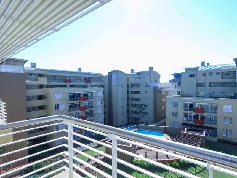 A spacious balcony featuring a metal balustrade, retractable awning, and an exterior air conditioning unit, overlooking the apartment complex grounds and pool area.