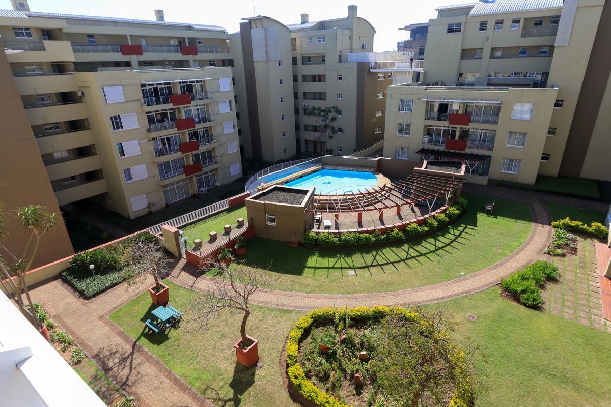 Aerial view of an apartment complex courtyard featuring a central swimming pool, landscaped gardens, paved pathways, and a pergola structure.