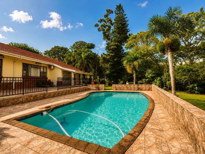 A well-maintained outdoor pool area featuring a tiled surround, brick retaining walls, and a metal security railing, set against a lush garden backdrop.