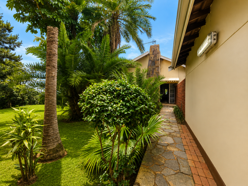 A well-maintained rear garden path featuring stone paving, lush tropical landscaping, and a distinctive stone chimney stack along the exterior wall.