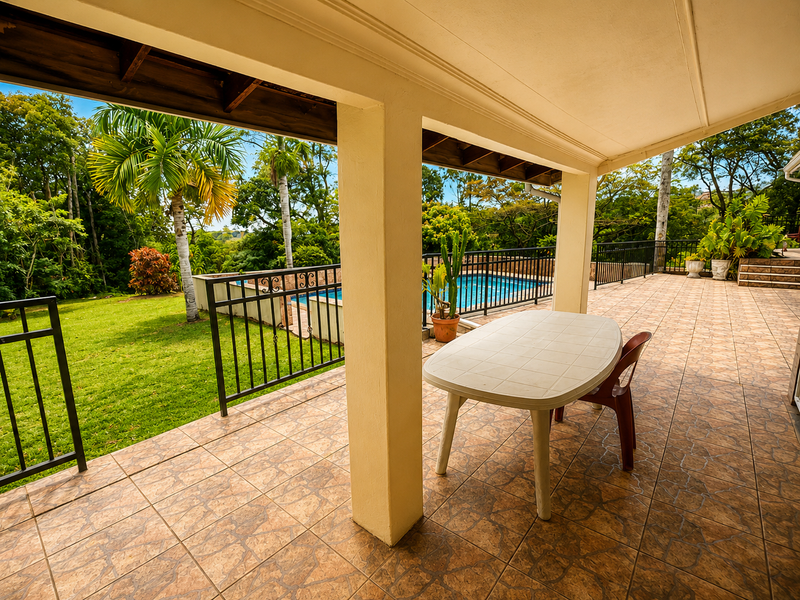 Spacious covered patio featuring tiled flooring and metal balustrades, overlooking a private swimming pool and lush garden area.