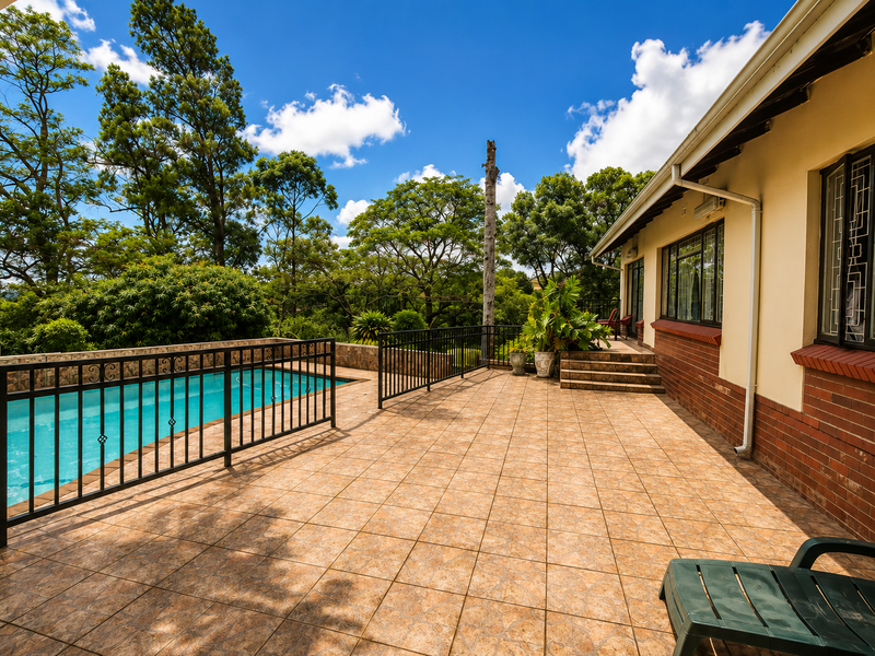 A spacious tiled patio area featuring a swimming pool with safety fencing, overlooking a lush garden setting.