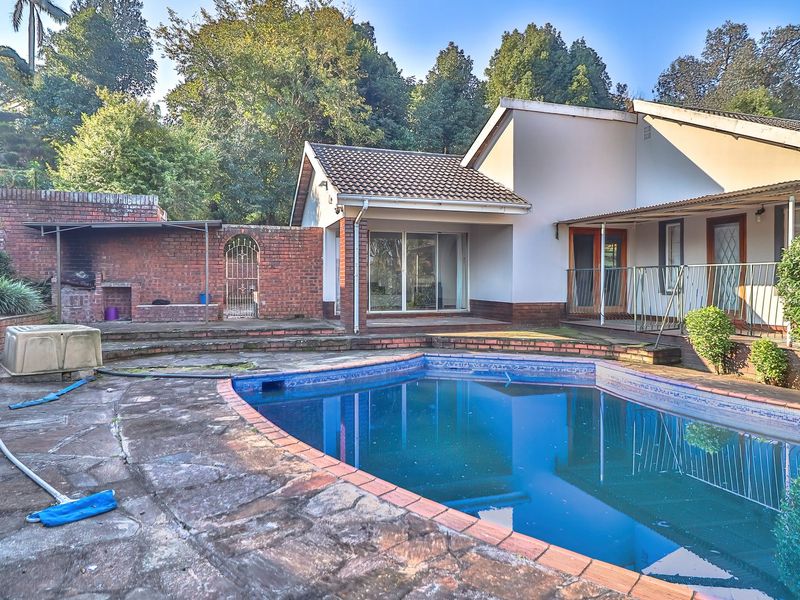 Outdoor pool area featuring a swimming pool, stone paving, and a brick built-in braai, set against a backdrop of mature trees.