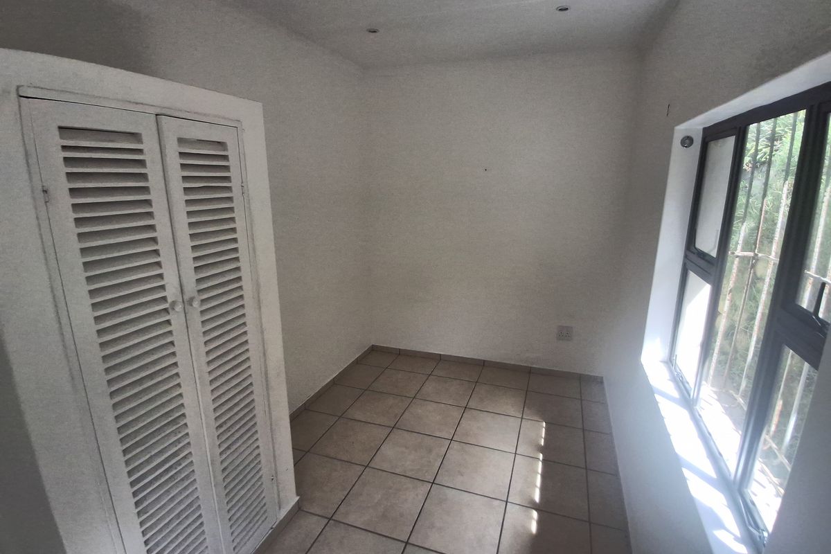 A compact, empty bedroom featuring tiled flooring, a built-in cupboard with louvered doors, and a window fitted with security bars.