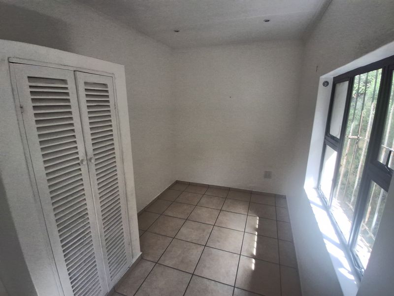 A compact, empty bedroom featuring tiled flooring, a built-in cupboard with louvered doors, and a window fitted with security bars.