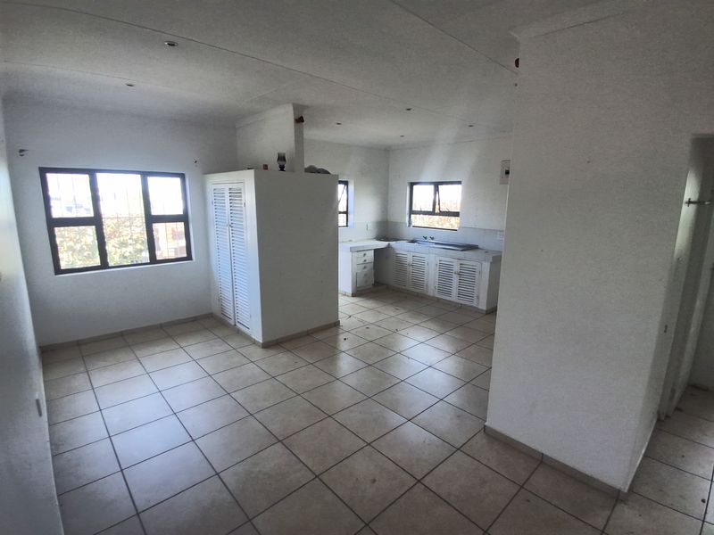 An open-plan living and kitchen area featuring tiled flooring, built-in cabinetry with louvered doors, and aluminium window frames in a neutral, vacant space.