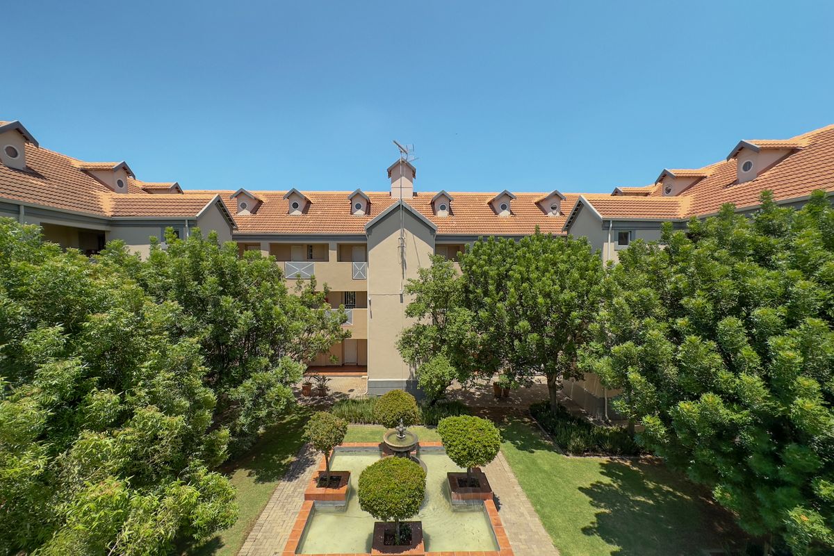 A well-maintained apartment complex courtyard featuring a central water fountain, landscaped gardens, and traditional terracotta roof tiles with dormer windows.