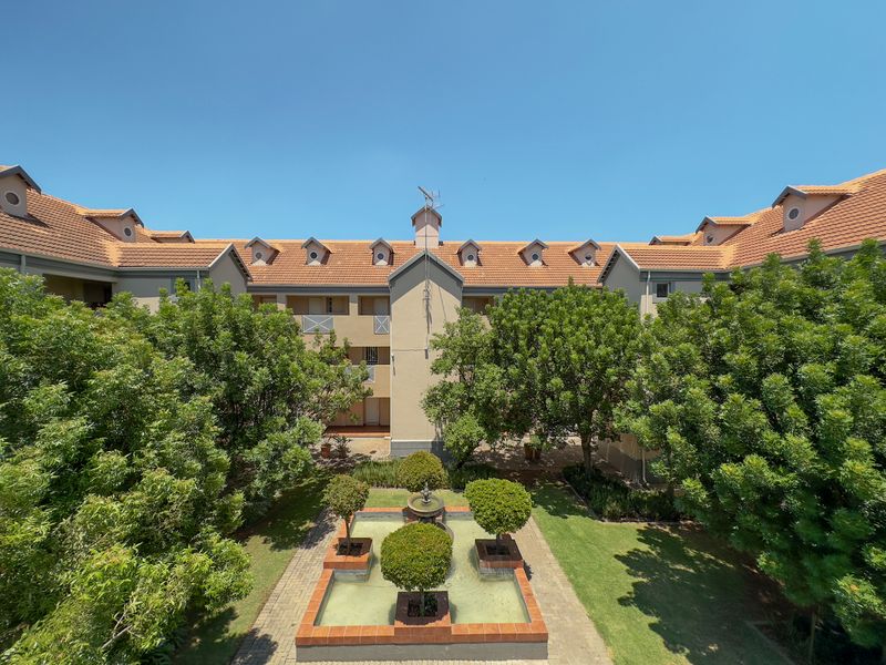 A well-maintained apartment complex courtyard featuring a central water fountain, landscaped gardens, and traditional terracotta roof tiles with dormer windows.